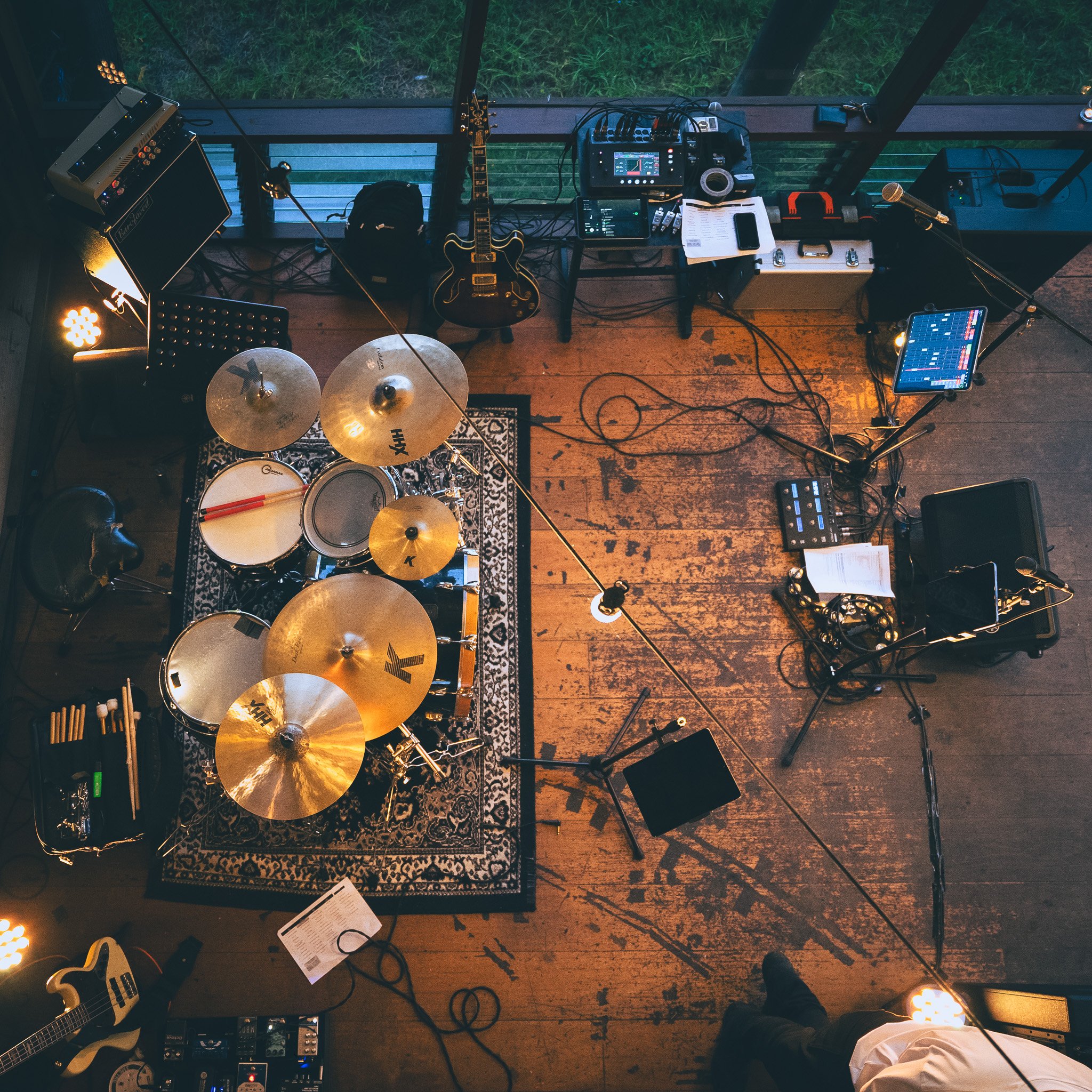 Elevated view of a music studio featuring a drum set on a patterned rug, multiple microphones, electronic equipment, guitars, and cables on a wooden floor with large windows showing greenery outside. Aubrey's set up before the dance floor gets going