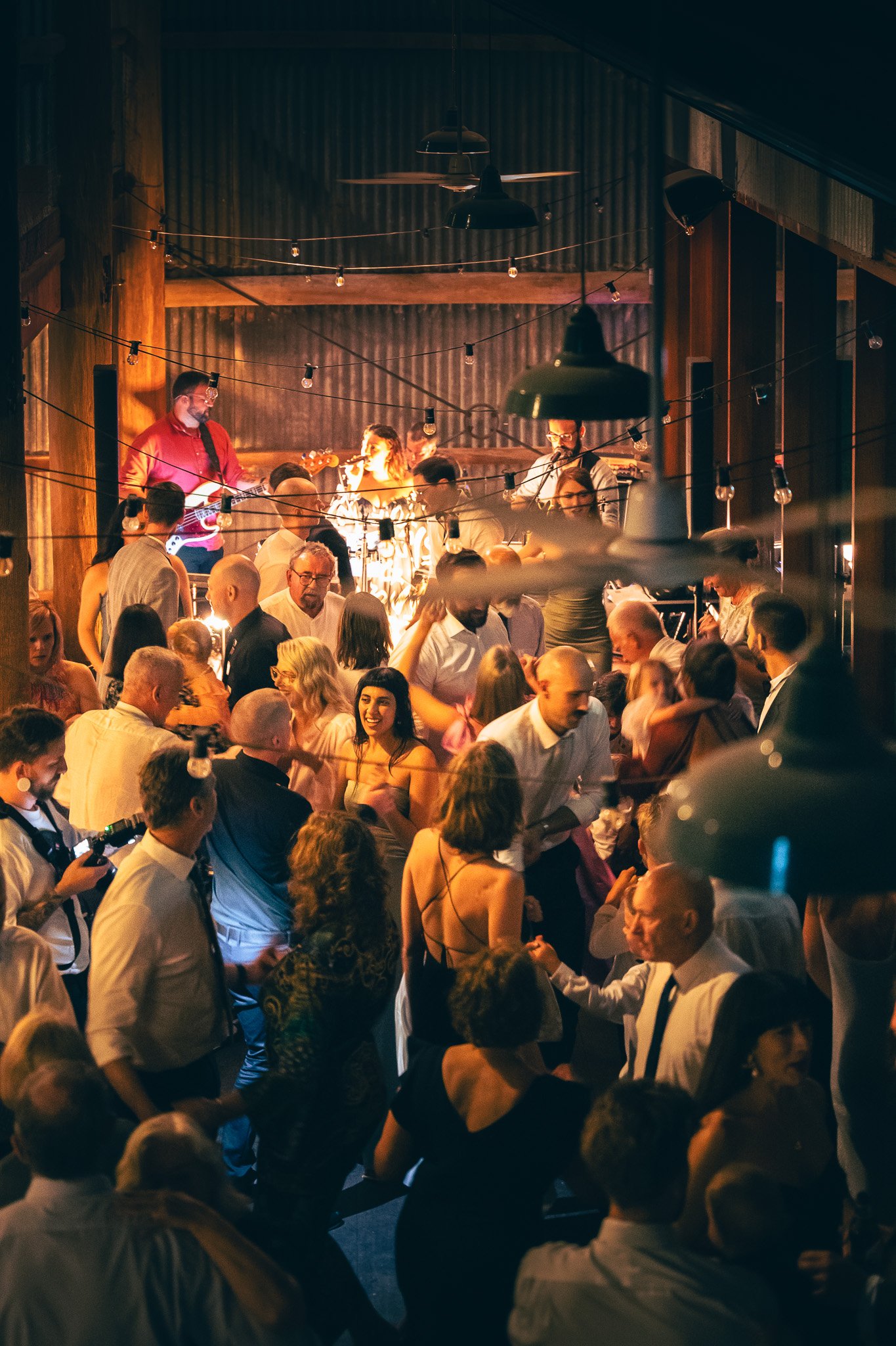 A lively indoor party with Aubrey, a live band, performing on a stage in Sydney, string lights hanging overhead, and many people dancing and socialising.