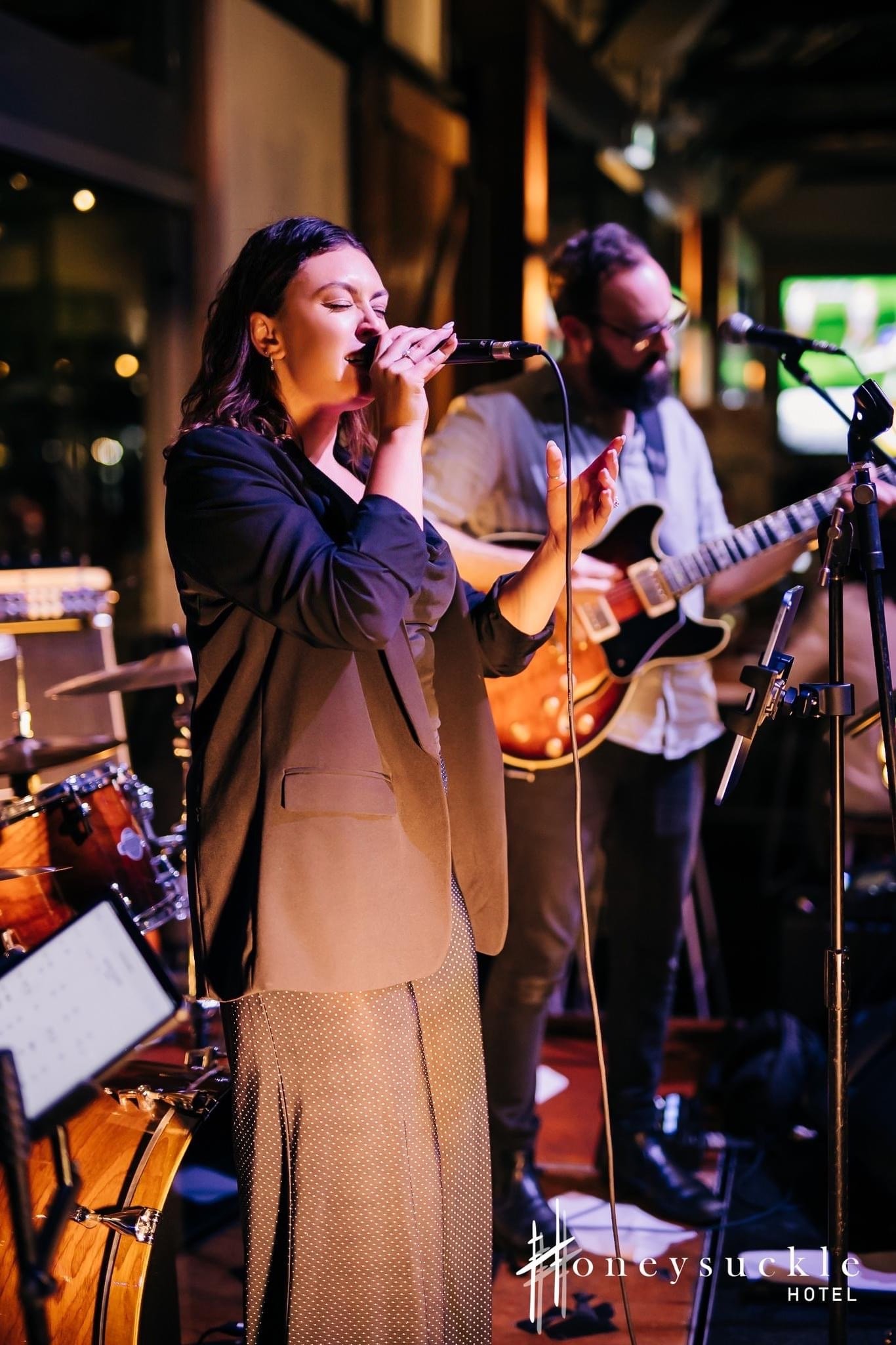 A woman singing into a microphone and a man playing an electric guitar during a live music performance at Honey Suckle Hotel.