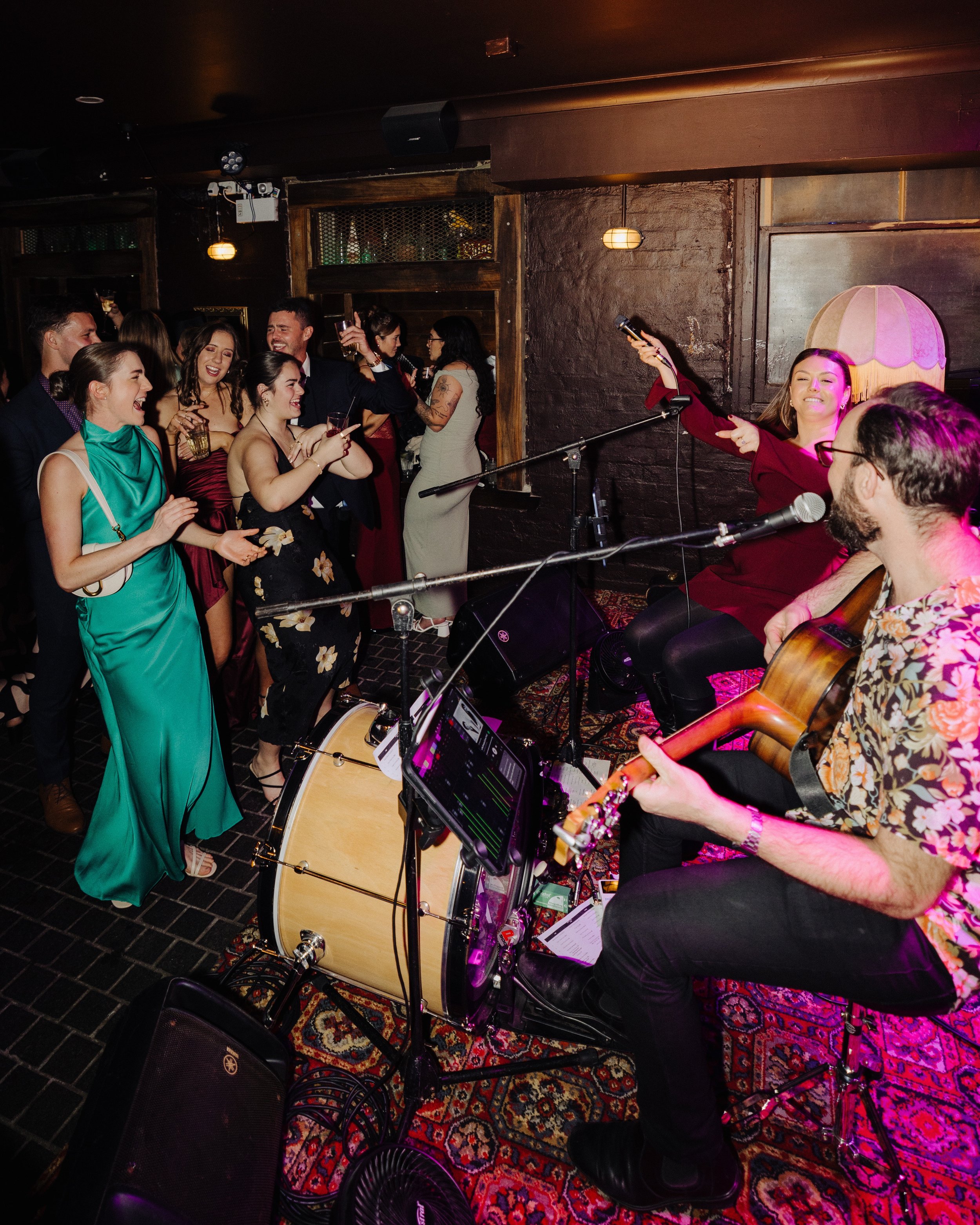 A lively party scene with India singing into a microphone while playing guitar, Jake playing a kick drum, and a group of people dancing and enjoying drinks at The Underground in Newcastle for a corporate networking event.